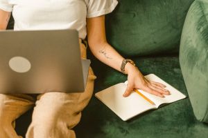Frau sitzt am Sofa mit Laptop auf den Beinen und hält ein Heft mit Bleistift geöffnet