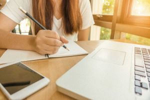 junge Frau schreibt mit Bleistift in einen Block davor ein Laptop
