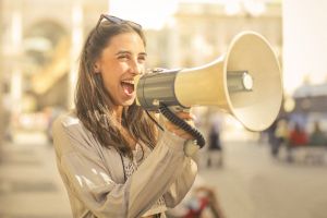 Junge Frau mit langen Haaren und Sonnenbrille in den Haaren spricht in ein Megaphon